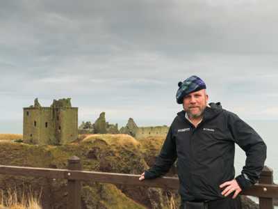 Highland Explorer Guide in a kilt and bonnet posing with the dramatic ruins of Dunnottar Castle perched on a coastal cliff
