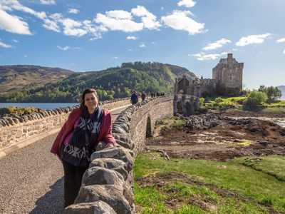 Woman standing on the bridge leading to Eilean Donan Castle, with the stone fortress and mountains in the background