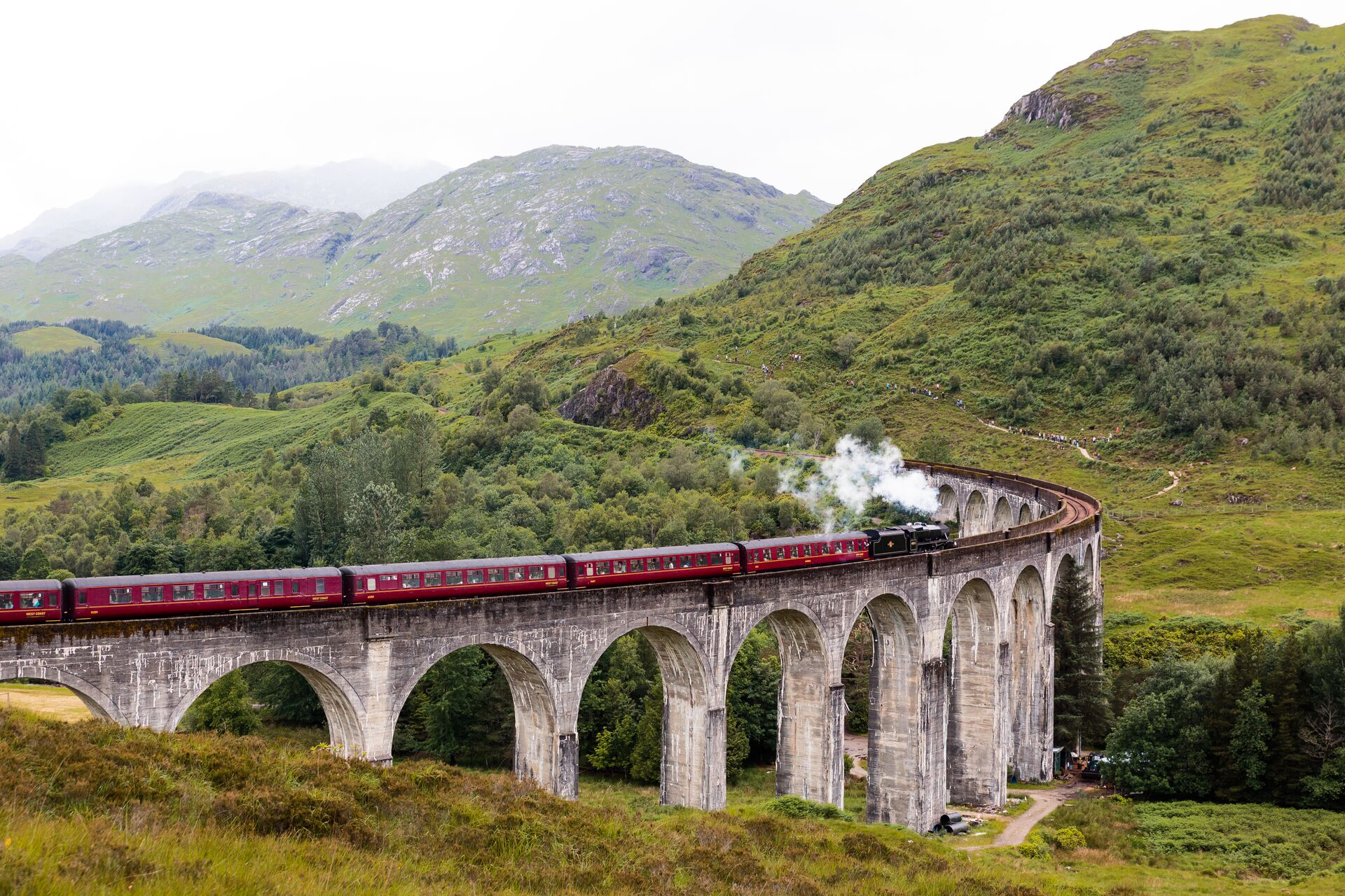Jacobite Steam Train: red train carriages and black steam engine crossing the iconic Glenfinnan Viaduct and lush hills in the background